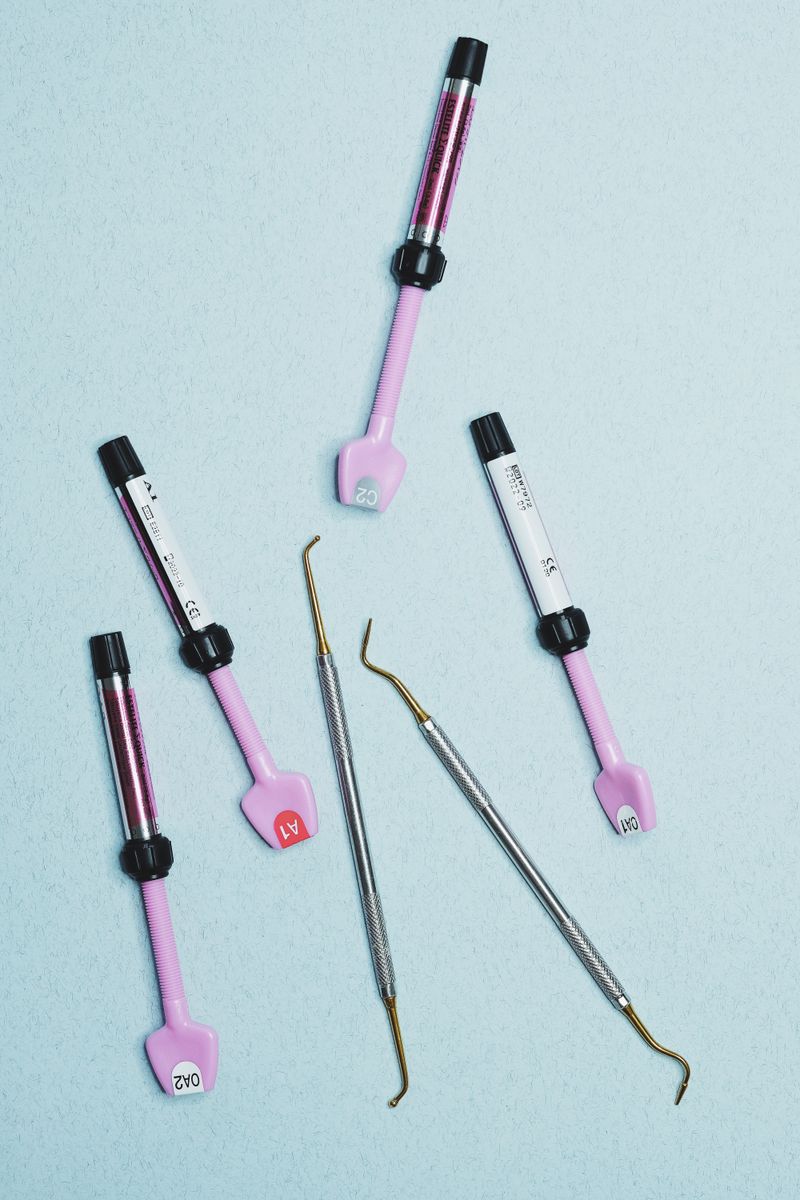 Five dental material syringes and two hand instruments laid on a pale blue surface